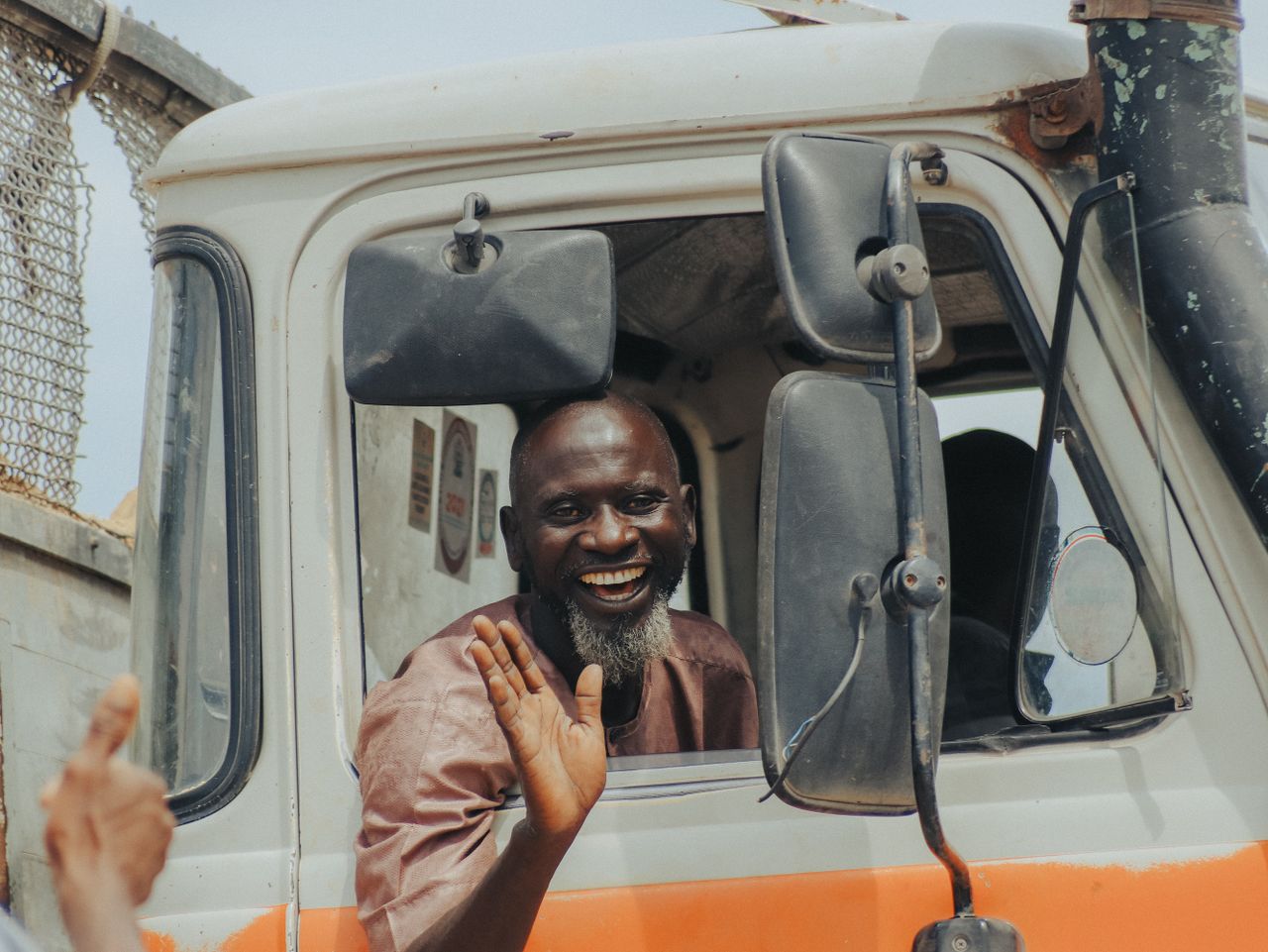 hero-img-01 Cheerful truck driver waving and smiling from his vehicle's window.