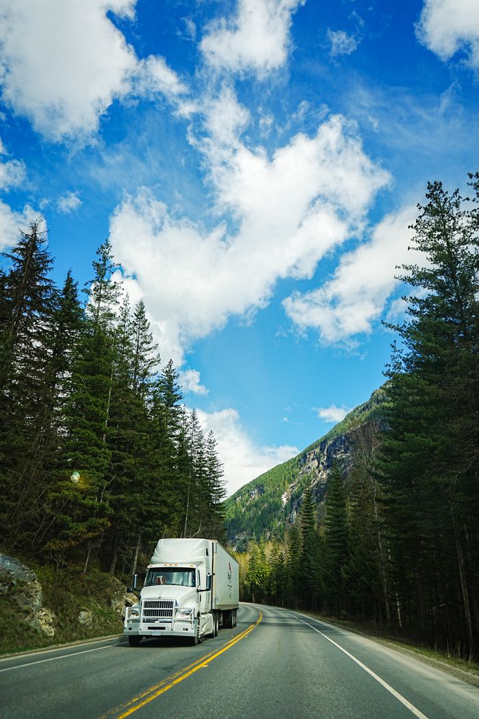 services-02 White delivery truck drives on a mountain road under a clear blue sky, surrounded by tall trees.