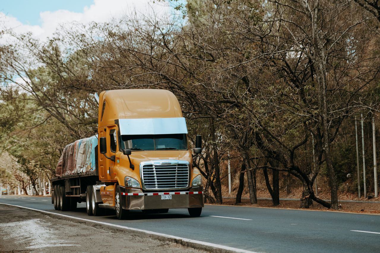 about-02 A yellow freight truck travels on a rural road lined with trees, capturing the essence of transport in El Salvador.